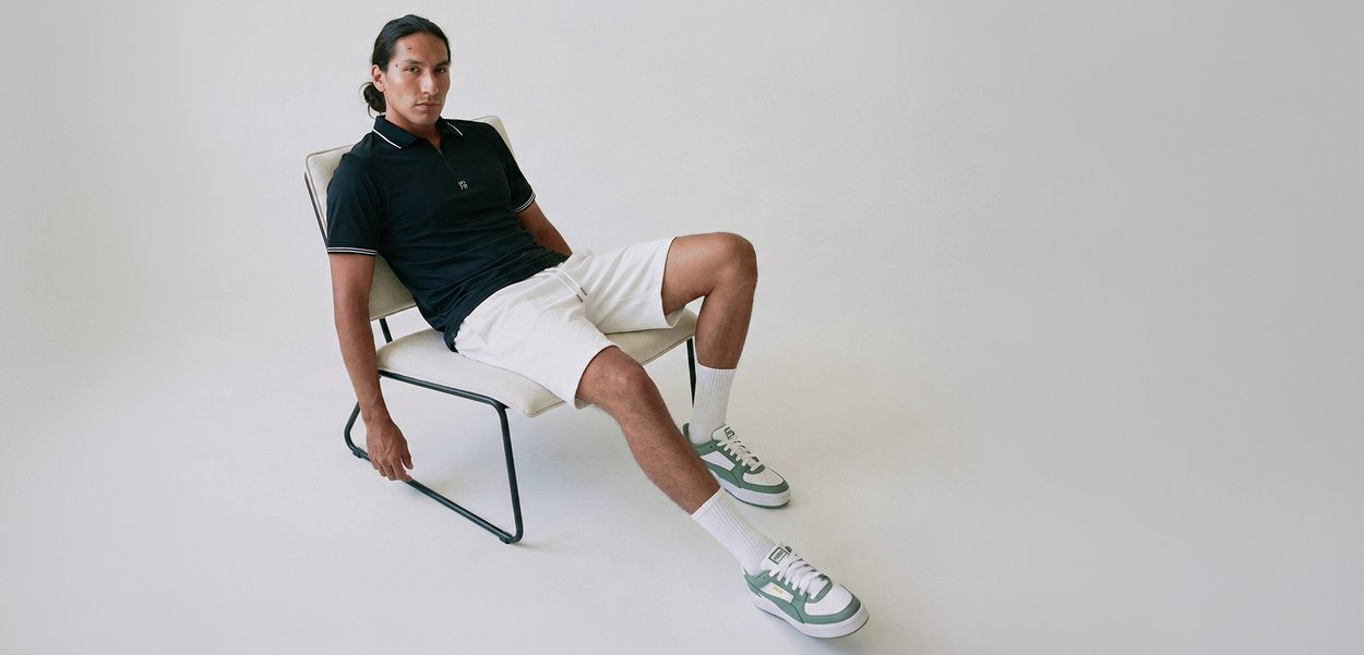 Nuori mies nojautuu minimalistisessa tuolissa, pukeutunut mustaan poolopaitaan, valkoisiin shortseihin, valkoisiin sukkiin ja vihreävalkoisiin tennareihin, yksinkertaisen taustan edessä.