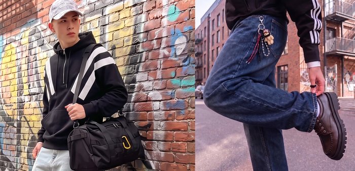 Person wearing dark jeans and brown shoes bending one leg to adjust the pant cuff on a street with brick buildings in the background.