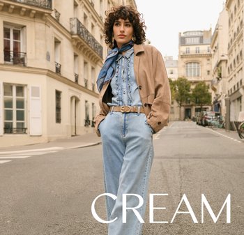 Frau trägt eine hellbraune Jacke, ein blaues Jeans-Hemd mit Knöpfen, hoch taillierte Jeans und einen blauen Schal und steht mit den Händen in den Taschen auf einer städtischen Straße.