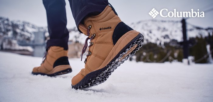 Nahaufnahme von beigefarbenen Columbia Winterstiefeln, die auf Schnee gehen, mit verschwommenen schneebedeckten Bergen im Hintergrund.