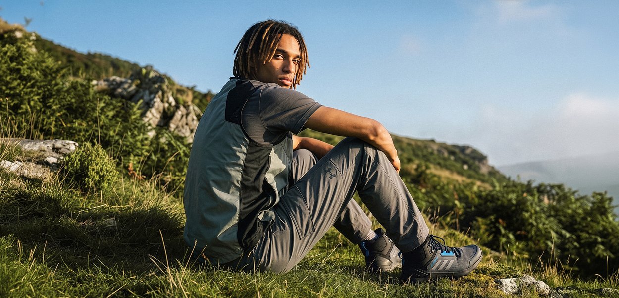 Jeune homme avec des dreadlocks portant des vêtements d'extérieur, assis sur l'herbe d'une colline avec un paysage rocheux et un ciel bleu en arrière-plan.