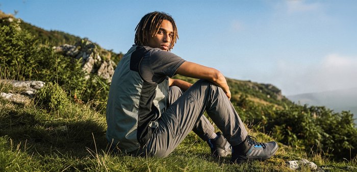 Jeune homme avec des dreadlocks portant des vêtements d'extérieur, assis sur l'herbe d'une colline avec un paysage rocheux et un ciel bleu en arrière-plan.