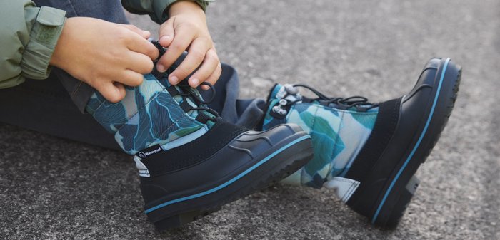 Kind sitzt auf dem Boden und schnürt blau-schwarz gemusterte Winterstiefel mit schwarzen Schnürsenkeln auf grauem Pflaster.