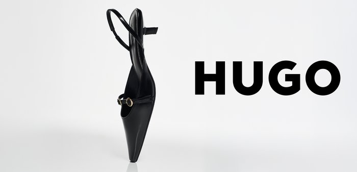 Zapato de tacón alto de punta negra con tira de hebilla y talón descubierto, colocado en posición vertical junto al logo audaz de HUGO en negro sobre fondo blanco.