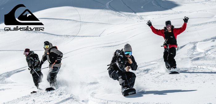Quatre snowboarders sur un terrain enneigé, portant des vestes colorées et des lunettes de protection. De la neige s'éjecte de leurs planches, avec en toile de fond des montagnes blanches.
