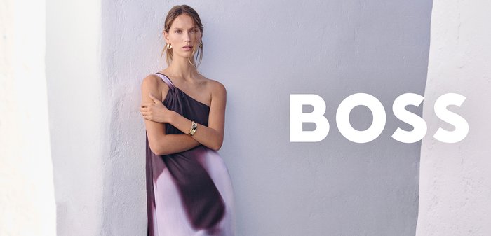 Woman wearing one-shoulder black and white gradient maxi dress, standing against a light textured wall with "BOSS" text.
