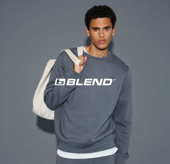 Felpa grigia con collo a giro e logo, realizzata in un tessuto liscio. Modello che tiene una borsa a tracolla beige, su uno sfondo semplice.