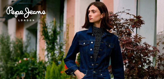 Sieviete tumši zilā džinsa kombinezonā ar daļēji kabatās ieliktiem rokām, stāv dārzkopības centrā, apkārt augu un koki.