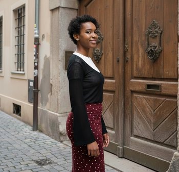 Νεαρή γυναίκα με κοντά σγουρά μαλλιά, που φοράει μαύρο τοπ και φούστα με βυσσινί πουά, στέκεται σε λιθόστρωτο δρόμο δίπλα σε μεγάλη ξύλινη πόρτα.