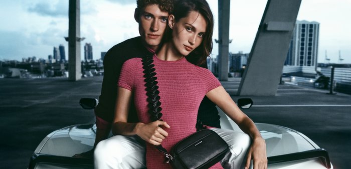 Jeune homme et femme assis sur une voiture blanche dans un parking souterrain, femme en robe rose tenant un sac bandoulière noir, silhouette de la ville en arrière-plan.