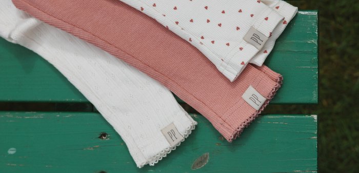 Three folded knitted baby leggings in white, pink, and white with red hearts, each with a small fabric tag on a green wooden surface.