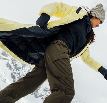Gelbe und schwarze puffy Jacke über einem schwarzen Oberteil, kombiniert mit braunen Cargo-Hosen. Dazu gehört eine strukturierte Mütze und Handschuhe, vor einer Kulisse aus schneebedeckten Bergen.