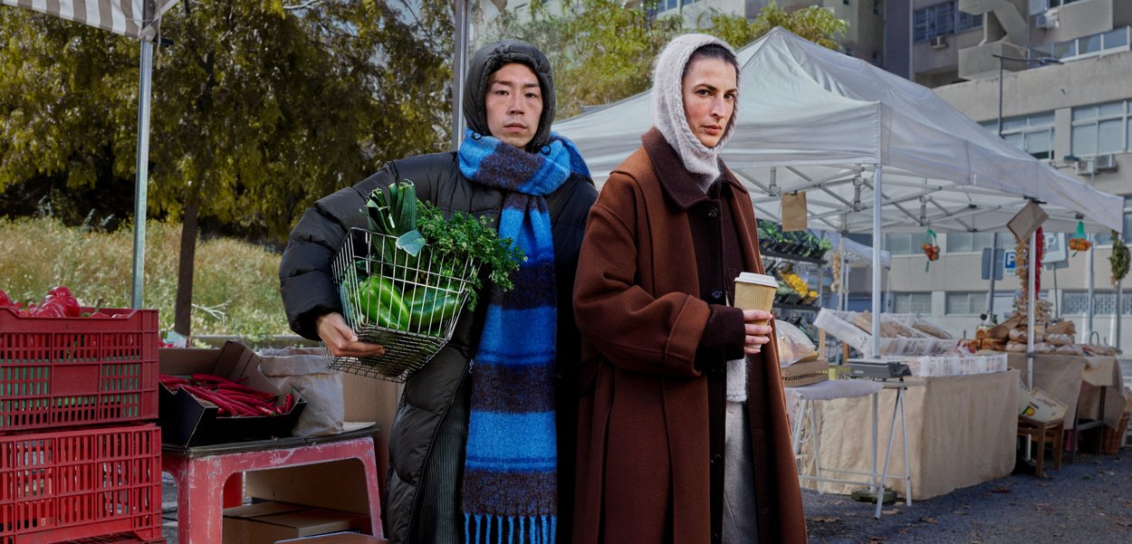 Two individuals stand in a market. One is holding a wire basket filled with greens, dressed in a black puffer jacket and a blue scarf. The other person is wearing a brown coat.