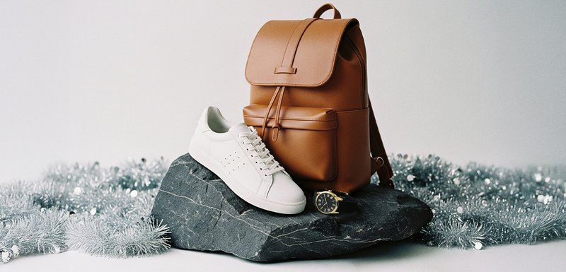 Καφέ δερμάτινος σακίδιο, λευκά αθλητικά παπούτσια και μαύρο ρολόι σε σκοτεινή πέτρινη επιφάνεια με ασημένιο γκλίτερ στο φόντο. Κλασικά στοιχεία σχεδίασης.