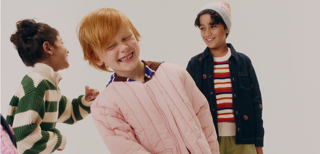 Trei copii râd împreună. Unul poartă o jachetă puffer roz deschis, altul un pulover cu dungi verzi și bej, iar al treilea o jachetă din denim.