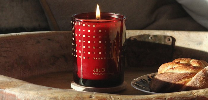 Bocal à bougie en verre rouge avec des motifs dorés, mèche allumée, reposant sur un sous-verre en bois, à côté d'une pâtisserie tressée sur une assiette.