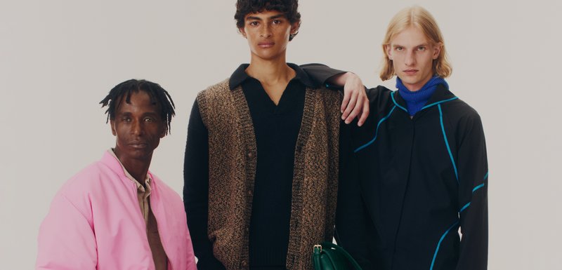 Trois modèles se tiennent ensemble. L'un porte une veste bomber rose, un autre un gilet texturé marron par-dessus une chemise noire, et le dernier un manteau noir avec des accents bleus.