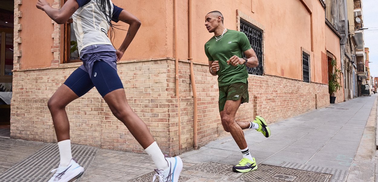 Dos personas corriendo por una acera de la ciudad junto a edificios de ladrillo, vistiendo ropa deportiva y zapatillas de correr.