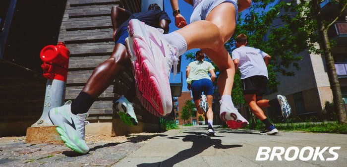 Cuatro personas corriendo por un camino urbano soleado, usando zapatillas deportivas, con un hidrante de incendios rojo y edificios al fondo.