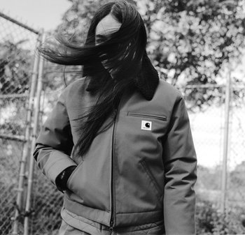 Chaqueta gris con cuello negro, bolsillos con cremallera y parche con logo, usada por una persona de cabello largo que sopla con el viento, de pie cerca de una valla.