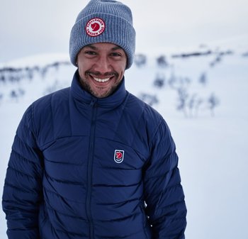 Navy Pufferjacke mit gestepptem Design und kleinem Logo auf der linken Brust. Hellgraue Strickmütze mit Logopatch. Schneereicher Hintergrund.
