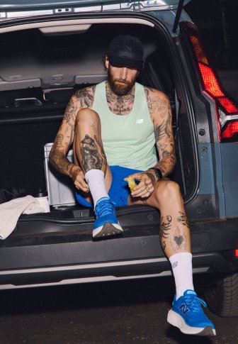 Un athlète en débardeur menthe et shorts bleus, portant des baskets bleues avec des accents blancs, assis dans le coffre d'une voiture, tenant une collation.