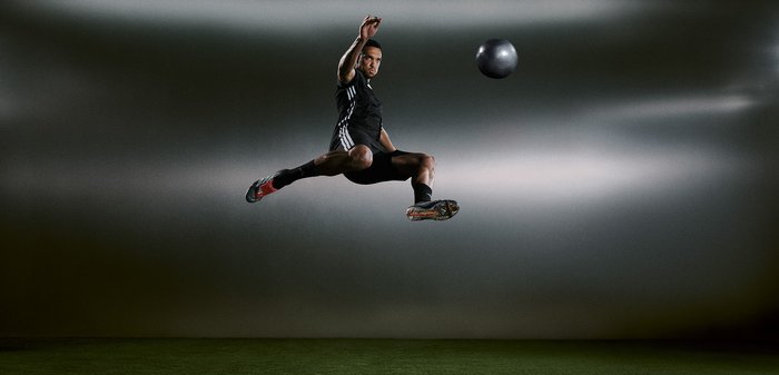 Un athlète en tenue de sport noire saute en l'air, donnant un coup de pied à un ballon gris. Ses chaussures présentent des accents colorés, avec une surface lisse et texturée.