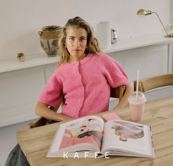 Rosa, kurzärmeliges, gestricktes Strickjacke mit Knöpfen und weicher Textur. Ein Holztisch trägt eine Zeitschrift und einen Smoothie in einem durchsichtigen Becher.