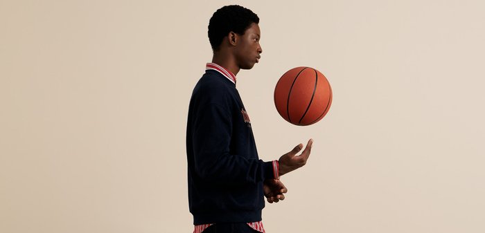 Sudadera de color navy con un cuello a rayas rojas y blancas, un balón de baloncesto flotando sobre una mano, contra un fondo neutro.