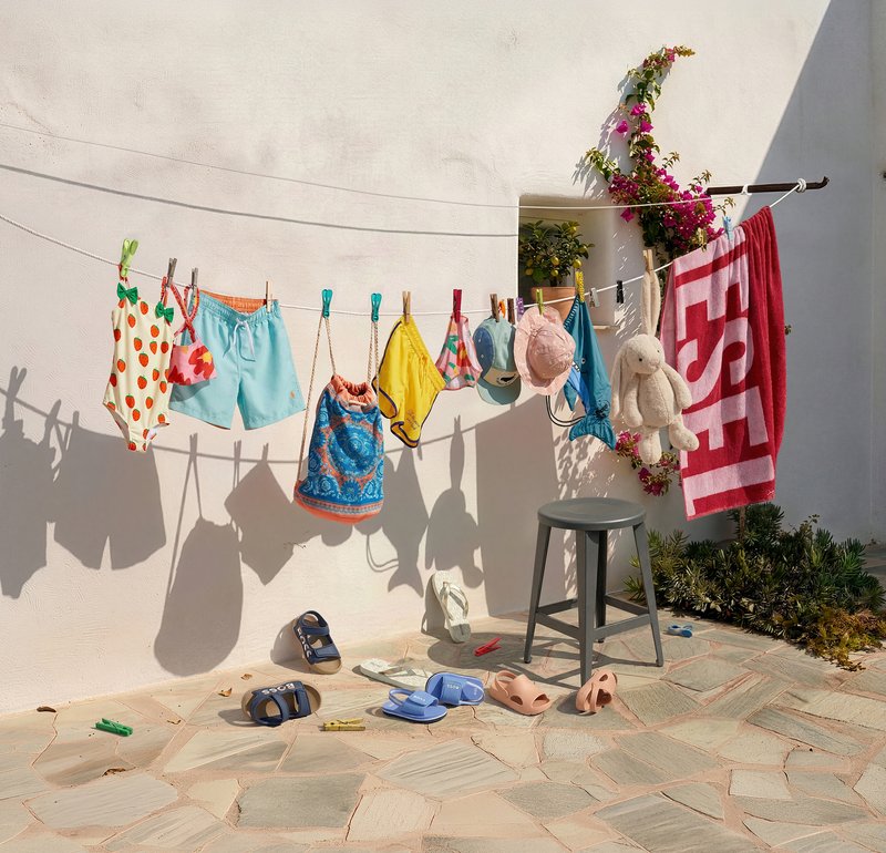 Ropa de baño infantil, toallas, sombreros y un conejito de peluche colgados en un tendedero junto con sandalias y un taburete en un patio de piedra.