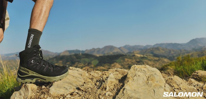 Leg wearing black and olive Salomon hiking boot and dark green Salomon sock stepping on rocky terrain with blurred hills in background.