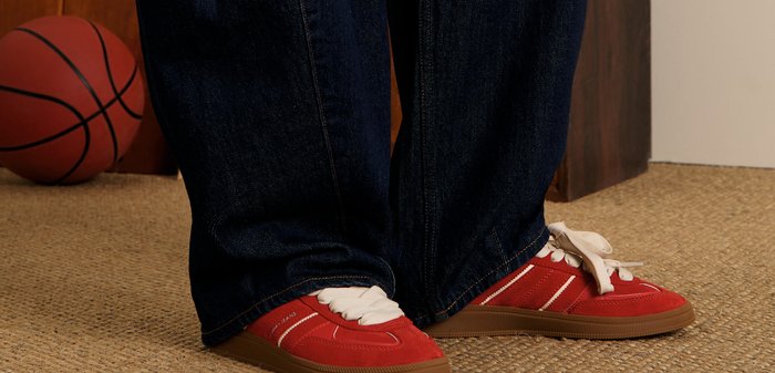Zapatillas de ante rojo con detalles en blanco y cordones beiges, combinadas con jeans de mezclilla azul oscuro. El fondo incluye un balón de baloncesto y una superficie texturizada.