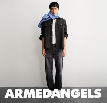 Schwarze Knopfjacket über einem weißen Hemd, kombiniert mit dunklen Jeans. Ein blau gestreifter Schal akzentuiert das Outfit. Weiße Sneakers runden den Look ab.