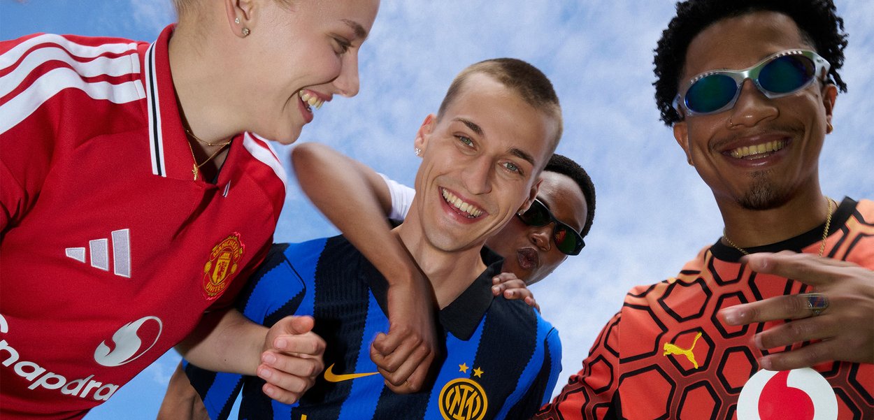 Cuatro jóvenes sonrientes que llevan diferentes camisetas de fútbol están juntos bajo un cielo azul, mostrando amistad y alegría.