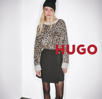 Frau mit schwarzer Mütze, Leopardmuster-Pullover, schwarzem Rock und Strumpfhosen, die an einer weißen Wand steht, mit "HUGO" Textüberlagerung.