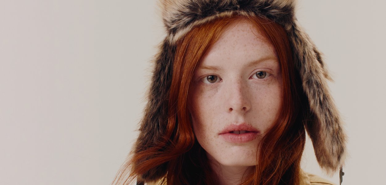 Căciulă din blană artificială cu clape pentru urechi; model maro și crem; textură moale; purtată cu părul roșu într-o formă liberă; fundal neutru.