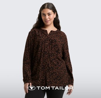 Kobieta ubrana w luźną brązowo-czarną koszulę z nadrukiem w panterkę na guziki i ciemne spodnie, stojąca na tle jasnoszarego tła.