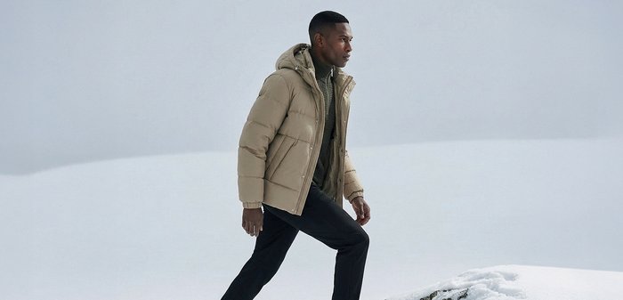 Doudoune beige avec capuche, dotée d'un motif matelassé et d'une fermeture éclair à l'avant. Portée par-dessus un pull vert foncé et assortie à un pantalon noir.