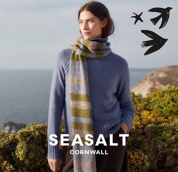 Modrý svetr s žebrovaným lemem, doplněný o vícebarevnou pruhovanou šálu v zelené a béžové barvě. Měkké textury s pobřežním pozadím.