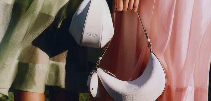 Zwei Personen in Pastellkleidern halten weiße Handtaschen mit einzigartigen geometrischen und sichelförmigen Mustern im Freien auf dem Gras.