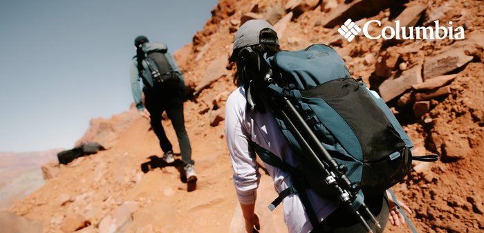 Columbia-Rucksack aus dunkelblauem und schwarzem Material mit externen Riemen, verstellbarem Tragesystem und Trekkingstöcken, getragen von Wanderern auf steinigem Gelände.