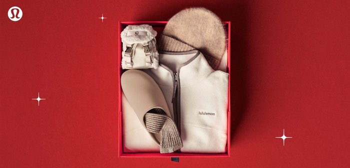 Rode cadeau doos met beige slippers, gebreide sokken, fleece jas, beanie en kleine donzige handtas op een rode achtergrond.