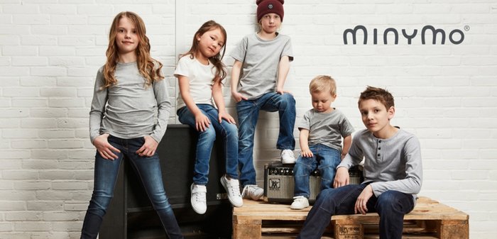 Vier Kinder posieren auf einer Holzleiter vor einer weißen Ziegelmauer. Sie tragen legere Kleidung, darunter graue und weiße Oberteile sowie Jeans.