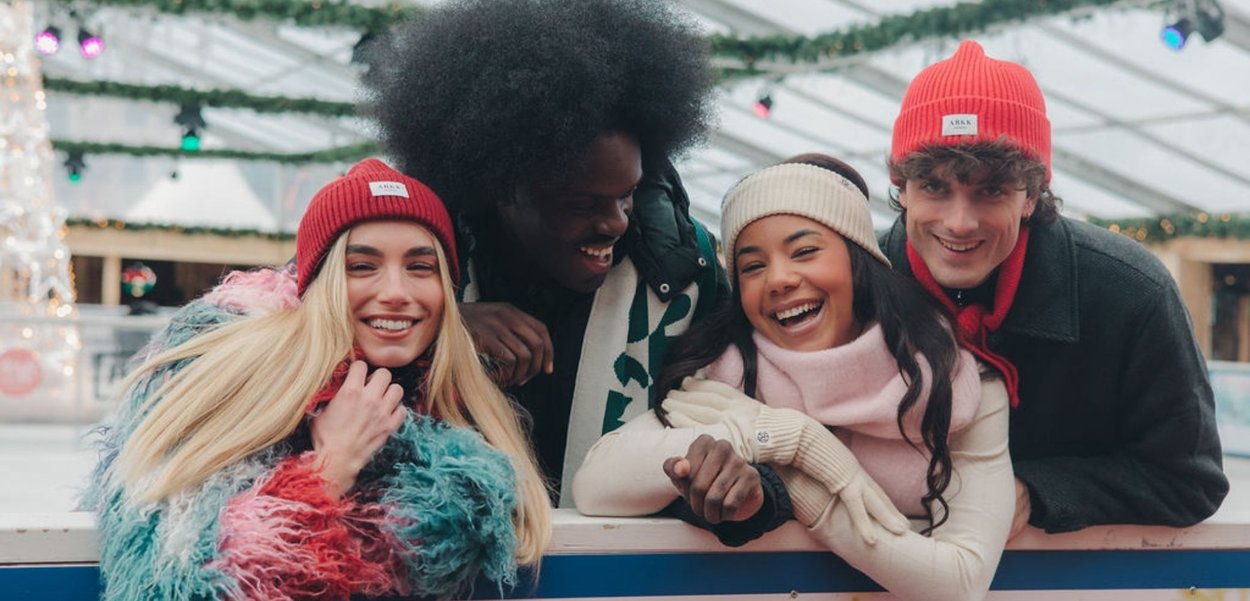 Vier personen lachen samen, gekleed in kleurrijke winterkleding: een roze en turquoise pluizige jas, een rode muts, een beige sjaal en handschoenen.