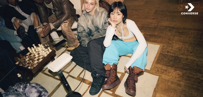 Două persoane stau pe un covor cu model. Poartă ținute casual; unul într-o jachetă gri metalizată și pantaloni negri, celălalt într-un top alb și pantaloni albaștri. Încălțămintea constă în adidași negri și maro cu gleznă înaltă.