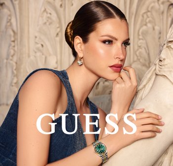 Orologio verde con dettagli in oro e argento, top senza maniche blu denim e orecchini decorativi. Modella posata accanto a una superficie scolpita.