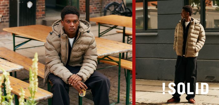 Beige Steppjacke mit Kapuze, ausgestattet mit einem Reißverschluss vorne und Seiten Taschen, getragen über einem schwarzen Shirt, kombiniert mit lockeren dunklen Hosen und Schuhen.