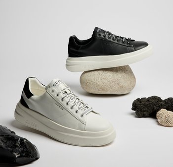 Sneakers in pelle bianca e nera con suole bianche spesse, dotate di lacci con marchio e accenti distintivi sul tallone. Posizionati su pietre texturizzate.