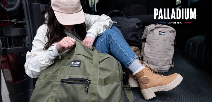 Zielona torba duffel z kieszenią na zamek i szczegółami paska; beżowe buty z teksturowymi podeszwami oraz niebieskie dżinsy, wszystko w pomieszczeniu.