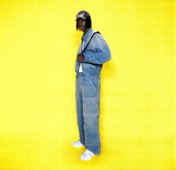 Giubbotto di jeans e pantaloni cargo in blu chiaro, abbinati a un cappellino con motivo e scarpe bianche. Lo sfondo è giallo uniforme.